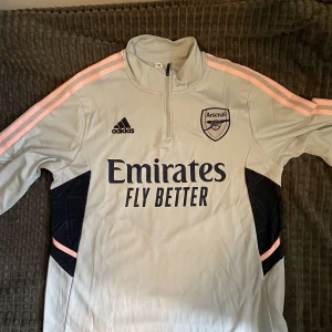 Arsenal Adidas träningströja ljusgrå - Snygg ljusgrå långärmad träningströja från Arsenal och Adidas med halv dragkedja. Tröjan har rosa ränder längs ärmarna och svarta detaljer på sidorna. Stort Arsenal-märke och Adidas-logga på bröstet. Tillverkad i funktionsmaterial som andas.