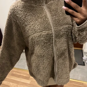 Beige teddyjacka/kofta med dragkedja - Mysig och oversized teddyjacka/ kofta i beige med hel dragkedja framtill. Jackan har en fluffig och mjuk textur, perfekt för kyliga dagar. Långärmad modell med hög krage som ger extra värme. Enkel och stilren design som är lätt att matcha.