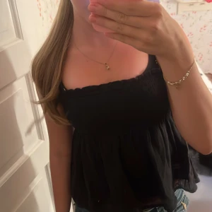 Svart linnetopp från Gina Tricot - Supersöt svart linnetopp från Gina Tricot med smockad överdel och tunna axelband som går att ta av. Toppen har en luftig, lätt utsvängd nederdel och volangkant upptill. Perfekt för varma dagar och enkel att styla med jeans eller kjol.