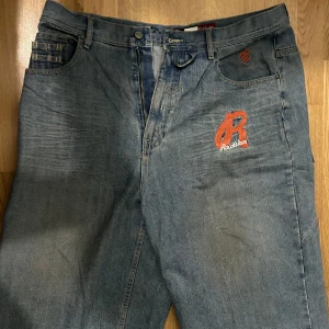 Rocawear jeans med brodyr och dragkedja - Säljer ett par blå jeans från Rocawear med röd broderad logga framtill och coola detaljer som extra fickor och dragkedja bak. Jeansen har en relaxed passform och klassisk femficksdesign. Perfekta för dig som gillar streetwear och unika detaljer.