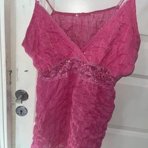 Rosa topp med spets och paljetter - Supersöt rosa topp med tunna axelband och v-ringad hals. Toppen har glittriga paljetter och pärlor längs med tyget och ett detaljerat spetsband under bysten. Materialet är lätt och skirt, perfekt för dig som gillar att sticka ut med färg och detaljer.