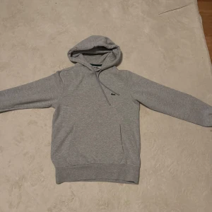Grå hoodie från Lacoste med huva - Snygg grå hoodie från Lacoste med klassisk krokodillogga på bröstet. Tröjan har huva med snörning, mjuk fleece på insidan och långa ärmar. Perfekt för chill dagar eller när du vill ha en clean och enkel stil.
