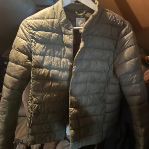 Olivgrön quiltad dunjacka - Säljer en olivgrön dunjacka med quiltad design och tryckknappar framtill. Jackan har en enkel, stilren look utan krage och är perfekt för kyliga dagar. Lätt och smidig att bära, passar till många outfits.Storlek 42 men är som en M/S