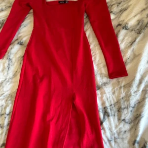 Röd långklänning från Boohoo - Säljer en snygg röd långklänning från Boohoo med fyrkantig urringning med en slits hela vägen upp till låren. Klänningen har en figurnära passform och är gjord i mjukt stretchigt material som sitter bekvämt. Perfekt för dig som vill sticka ut med en enkel men stilren look.