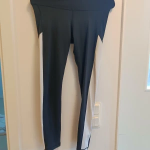 Svarta och vita leggings från Reebok - Snygga svarta leggings från Reebok med vita paneler längs sidorna och stort Reebok-tryck på benet. Tight passform och stretchigt material som passar perfekt till träning eller chill. Sportig look och bekväma att bära hela dagen.