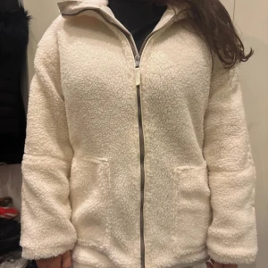 Vit teddyjacka med dragkedja - Mysig vit teddyjacka med dragkedja framtill och två stora fickor. Jackan har en hög krage och en cool detalj med en ljus rand längs ärmarna. Perfekt för kalla dagar när du vill vara både varm och snygg. Oversized passform för en chill vibe.