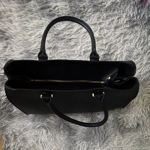 Svart stilren handväska i skinn - Snygg svart handväska med två rejäla handtag och guldfärgade metalldetaljer, och en dragkedja i mitten för extra säkerhet. Perfekt för dig som vill ha en klassisk och rymlig väska med stilren look. Köpt från hm länge sen❣️