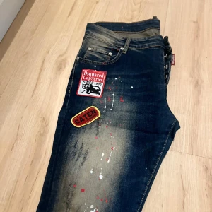 Dsquared2 jeans med patchar & färgstänk - Mörkblå jeans från Dsquared2 med coola patchar på framsidan och färgstänk i rött, vitt och gult. Jeansen har klassisk femficksdesign, slitna detaljer och Dsquared2-logga bak. Snygg streetstil med unik look.