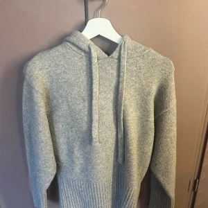 Grå stickad hoodie från Zara - Mysig grå hoodie från Zara i stickad material med huva och snörning. Tröjan har långa ärmar och breda ribbade muddar vid nederkant och ärmslut. Perfekt för dig som gillar chill och enkel stil med lite extra värme.