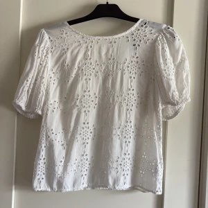 Vit broderad blus från KappAhl - Superfin vit blus från KappAhl i bomull med broderade hålmönster och puffiga korta ärmar. Blusen har v-ringning framtill och elastiska ärmslut för en söt och luftig känsla. Perfekt för en fräsch och somrig look!