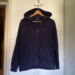 Mörkblå hoodie från Polo Ralph Lauren - Snygg mörkblå hoodie från Polo Ralph Lauren med röd logga på bröstet. Tröjan har huva, dragkedja framtill och två stora fickor. Tillverkad i mjuk bomull, perfekt för chill dagar. Klassisk och enkel design som alltid funkar.