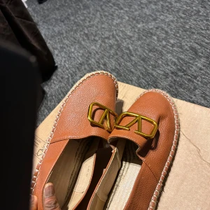 Valentino Garavani bruna loafers - Snygga loafers från Valentino Garavani i brun skinn med grov struktur. Dekorerade med en stor guldfärgad metall-logga framtill och har en flätad kant runt sulan. Platt sula och rund tå gör dem både stilrena och bekväma. Perfekt för dig som gillar lyxiga detaljer.