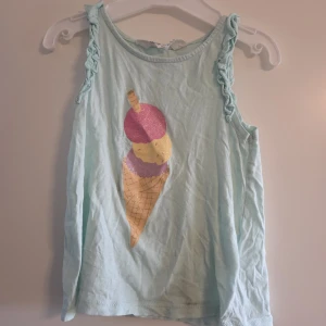 Ljusblå ärmlös topp med glasstryck H&M - Söt ljusblå/turkos linnetopp från H&M med glittrigt glassmotiv framtill i rosa, lila och gul. Toppen har volangdetaljer längs axelpartiet och är tillverkad i mjuk 100% bomull. Perfekt för sommarens alla äventyr!