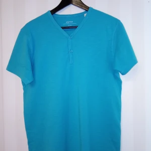 Turkos t-shirt med knappar från Esprit - Snygg turkos t-shirt från Esprit med v-ringning och två knappar framtill. Modellen är kortärmad och i 100% bomull, vilket gör den riktigt skön och luftig. Perfekt för dig som gillar en enkel men stilren look.                    Kan skickas med extra pris via post.