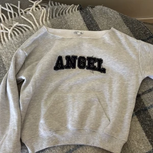 Grå sweatshirt Angel Subdued XS - Grå sweatshirt från Subdued i storlek XS med svart text 'ANGEL' framtill och stora svarta änglavingar på ryggen. Tröjan har bred halsringning, ribbade muddar och är tillverkad i mjuk bomullsblandning. Perfekt för dig som gillar statement-plagg. Orginal pris=574kr