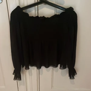 Svart offshoulder blus Gina Tricot - Svart offshoulder blus från Gina Tricot i transparent, lätt material. Vida långa ärmar med smock och volangdetalj vid ärmslut. Smockad rygg för skön passform och flowig nederkant. Perfekt för dig som gillar trendiga och feminina plagg.