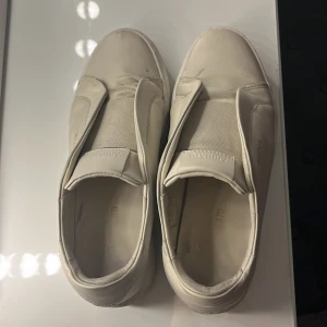 Vita sneakers från Axel Arigato -  vita sneakers från Axel Arigato i skinn med elastiskt band över vristen istället för klassisk snörning. Diskret logga i guld på sidan och på plösen. Platt sula och rund tå ger en clean look som passar till allt. Pris kan diskuteras 