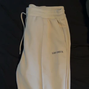 Beige mjukisbyxor från Les Deux - Snygga beige mjukisbyxor från Les Deux med broderad logga på benet. Byxorna har snörning i midjan och är tillverkade i mjukt material som ger en chill och avslappnad vibe. Perfekta för att hänga med kompisar eller bara ta det lugnt hemma.