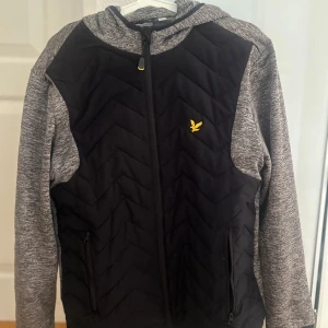Svart och grå pufferjacka Lyle & Scott - Snygg pufferjacka från Lyle & Scott i svart och grått med huva och dragkedja framtill. Jackan har quiltad front, gråmelerade ärmar och huva, samt två fickor med dragkedja. Gult Lyle & Scott-logo på bröstet ger extra stilpoäng. Två innerfickor på båda sidorna!