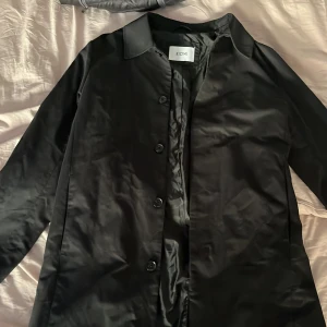 Svart rock från KIOMI - Snygg svart rock från KIOMI med klassisk krage och knappar framtill. Jackan har en stilren och rak passform, perfekt för dig som gillar minimalistisk stil. Materialet är slätt och har en lätt glansig finish.