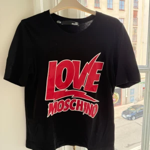 Svart Love Moschino t-shirt med tryck - Svart t-shirt från Love Moschino med stort rött och vitt tryck på framsidan. Klassisk rund hals och korta ärmar. Materialet är mjuk bomull som känns skönt mot huden. Passformen är normal och designen är clean med fokus på det grafiska trycket.