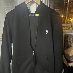 Svart hoodie från Polo Ralph Lauren - Svart hoodie från Polo Ralph Lauren med klassisk vit broderad logga på bröstet. Tröjan har dragkedja framtill, huva och två fickor. Tillverkad i mjuk bomull och har en clean, stilren look som funkar till allt. Perfekt för dig som gillar streetwear och enkel stil.