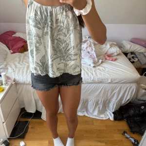 Ljusgrå linnetopp med palmtryck - Superfin ljusgrå linnetopp med tunna axelband och tropiskt palmbladsmönster. Toppen är luftig och har en lös passform, perfekt för varma dagar. Snygg detalj med extra band i ryggen som ger en cool look. Passar perfekt till shorts eller kjol!