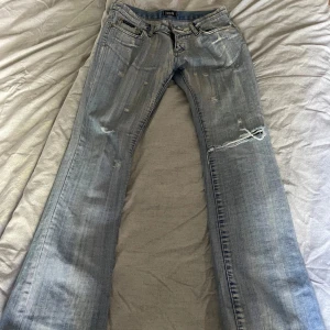 Ljusblå slitna jeans från D&G - Säljer ett par ljusblå jeans från D&G med slitningar och trasiga detaljer, särskilt vid knät. Klassisk femficksmodell med broderad logga på bakfickan. Jeansen har raka ben och är tillverkade i denim med en snygg tvättad look.