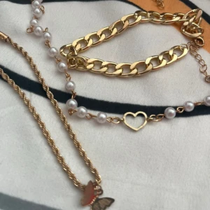 Guldiga smycken med pärlor och fjäril - Säljer ett set med smycken i guldton: ett chunky armband med breda länkar, ett halsband med tvinnad kedja och en röd fjärilshänge, samt ett pärlarmband med vita pärlor och ett hjärtformat guldfäste. Perfekt för dig som gillar detaljer och mixade stilar.