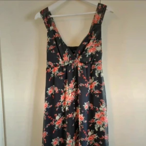 Klänning VILA strl M - En klänning i fint skick. Klänningen har svart botten och blommor och blad går i rosa, röda och gröna nyanser.  Modellen är kortare modell knälång cirka 100 cm lång. Klänningen har två lager ett tunt och skirt ytter tyg med blommorna och en fast skydd underklänning i svart, har även en liten dragkedja i sidan av klänningen, se bilder. INFO: Hund i hemmet.