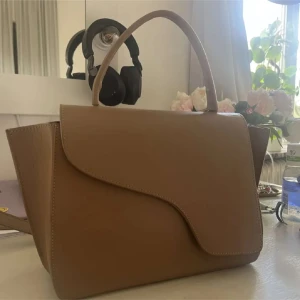 ATP väska  - Stilren beige axelväska i skinn med cool asymmetrisk lockdesign och både handtag och avtagbar axelrem. Väskan har guldfärgade metalldetaljer och rymligt huvudfack, perfekt för att bära med sig allt viktigt. Enkel att matcha och ger en clean look. Använd men bra skick 