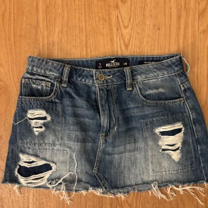 Blå jeanskjol från Hollister - Säljer en blå jeanskjol från Hollister med hög midja och slitna detaljer med sitter som low eaist. Kjolen har fransig nederkant, klassiska fickor fram och bak samt coola hål och slitningar för en edgy vibe. Materialet är 100% bomull och passformen är normal.