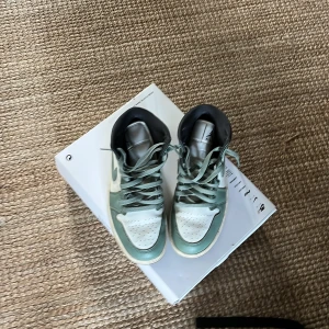 Nike Air Jordan 1 Mid Sage Green 40 - Nike Air Jordan 1 Mid i Sage Green och vitt skinn med klassisk swoosh och snörning. Snygg kontrast mellan de gröna och vita panelerna, svart detalj vid häl och platt sula. Perfekta för dig som vill sticka ut med en ikonisk sneaker.