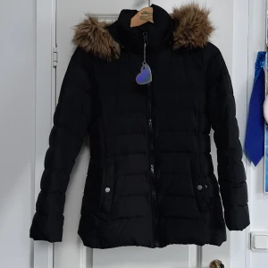 Svart pufferjacka Tommy Hilfiger S - Svart vinterjacka från Tommy Hilfiger i storlek S. Jackan har huva med fejkpäls, dragkedja framtill och två fickor med tryckknappar. Perfekt för kalla dagar och har en klassisk, stilren look.
