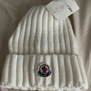 Vit ribbstickad mössa från Moncler - Vit ribbstickad mössa från Moncler med klassisk uppvikt kant och broderad logotyp framtill. Tjockt stickad i mjukt material som håller dig varm och ger en clean look. Perfekt accessoar för kyliga dagar.
