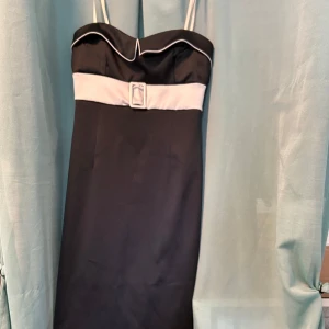 Svart medellångklänning med vit detalj - Snygg svart klänning med vita axelband och vit bård över bysten, prydd med ett dekorativt bälte framtill. Klänningen har en stilren, figurnära passform och med en elegant kant upptill. Perfekt för dig som gillar klassisk och chic stil.