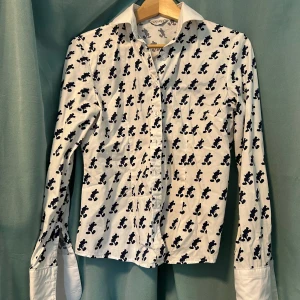 Vit skjorta med Mickey Mouse-mönster - Vit skjorta med svarta Mickey Mouse-silhuetter över hela tyget. Klassisk krage, långa ärmar och knäppning framtill. Manschetterna är vita med blå broderade initialer 'D.K.'. Skjortan är i bomull och har en lekfull men stilren vibe.
