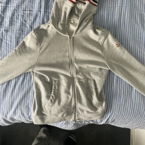 Grå Moncler hoodie med dragkedja - Snygg grå hoodie från Moncler med hel dragkedja och huva. Tröjan har Moncler-logga på ärmen och coola ränder i rött, vitt och blått på insidan av huvan. Mjuk bomullskänsla och två fickor framtill. Perfekt för chill eller streetwear.