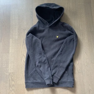 Svart hoodie från Lyle & Scott - Svart hoodie från Lyle & Scott med klassisk gul logga broderad på bröstet. Tröjan har huva, känguruficka och ribbade muddar. Perfekt för en chill och stilren look. Tillverkad i mjuk bomull för extra komfort.