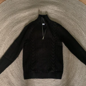 Svart stickad half zip från Jack & Jones - Säljer en svart stickad tröja från Jack & Jones i storlek XS. Tröjan har ett snyggt kabelstickat mönster, hög krage och en half zip-dragkedja framtill. Perfekt för dig som gillar stilren och enkel design med en twist.
