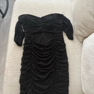 Svart omlott kortklänning med rynk - Snygg svart kortklänning med omlott-design och rynkade detaljer längs hela klänningen. Offshoulder och med långa ärmar ger en cool och trendig vibe. Perfekt för dig som gillar att sticka ut med stilrena detaljer.