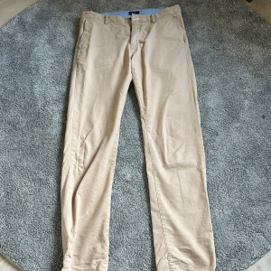 Beiga chinos från Gant, stl 176 - Snygga beiga chinos från Gant i klassisk rak modell. Byxorna har bälteshällor, sidofickor och bakfickor med knapp. Tillverkade i mjuk bomull som känns skön mot huden. Perfekta för en clean och stilren look. Använde några gånger och är i bra skick. En liten ”fläck” på högra benet nertill. Går att justera i midjan.