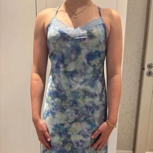 Zara klänning - Säljer en långklänning i mjukt, glansigt material med blått, lila och grönt blommigt mönster. Klänningen har tunna axelband, spetsdetaljer vid byst och rygg samt öppen rygg med korsade band. Dragkedja bak för enkel påklädning. Perfekt för sommarens alla tillfällen!