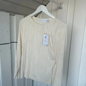 Krämvit ribbad topp från Vila - Supersnygg krämvit långärmad topp från Vila med vågigt ribbat mönster. Toppen har rund halsringning och är tillverkad i ett mjukt, stretchigt material som sitter skönt på kroppen. Perfekt att styla med jeans eller kjol för en clean look.