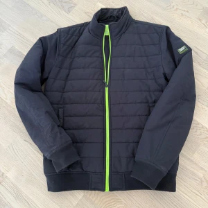 Svart pufferjacka med grön dragkedja - Svart pufferjacka från SuperDry med en snygg neongrön dragkedja framtill. Jackan har ståkrage, quiltad design och ribbade muddar vid ärmslut och nederkant. Perfekt för dig som vill ha en stilren jacka med en cool detalj. Nypris 1200kr, Storlek L men passar som S.