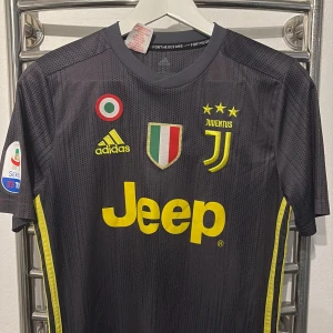 Juventus Ronaldo 7 Adidas fotbollströja - Juventus bortatröja med Ronaldo 7 på ryggen. Svart med gula detaljer, Juventus-logga, tre stjärnor, Adidas-märke och Jeep som sponsor. Korta ärmar och tillverkad i lätt, ventilerande material. Perfekt för dig som älskar fotboll och vill sticka ut på planen.