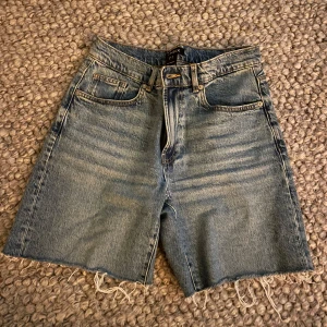 Blå jeansshorts med rå kant från Lindex - Snygga blå jeansshorts från Lindex i klassisk modell med fem fickor och rå, fransig kant nertill. Shortsen har normal passform och midja, perfekt för en chill sommarlook. Tillverkade i slitstarkt denimtyg med cool tvättad effekt.