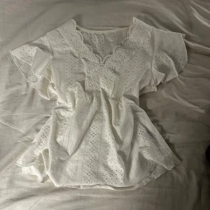 Vit blus med spets och broderi - Superfin vit blus med v-ringning, spetsdetaljer runt halsen och ärmarna samt broderade hålmönster över hela tyget. Blusen har korta, luftiga ärmar och är lätt utsvängd nedtill.