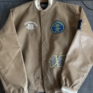 Beige bomberjacka BoohooMAN med patches - Snygg beige bomberjacka från BoohooMAN med coola patches och broderade detaljer. Jackan har kontrasterande ärmar i skinnimitation, tryckknappar och ribbade muddar. Perfekt för dig som vill sticka ut med en streetinspirerad look.