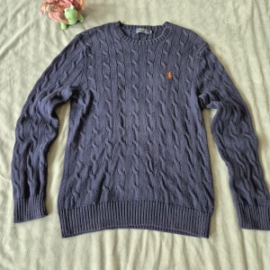Mörkblå stickad tröja Polo Ralph Lauren - Kabelstickad mörkblå tröja från Polo Ralph Lauren med klassisk orange logga broderad på bröstet. Tröjan har rund halsringning, ribbade muddar och är långärmad. Perfekt för dig som gillar en tidlös och clean stil.